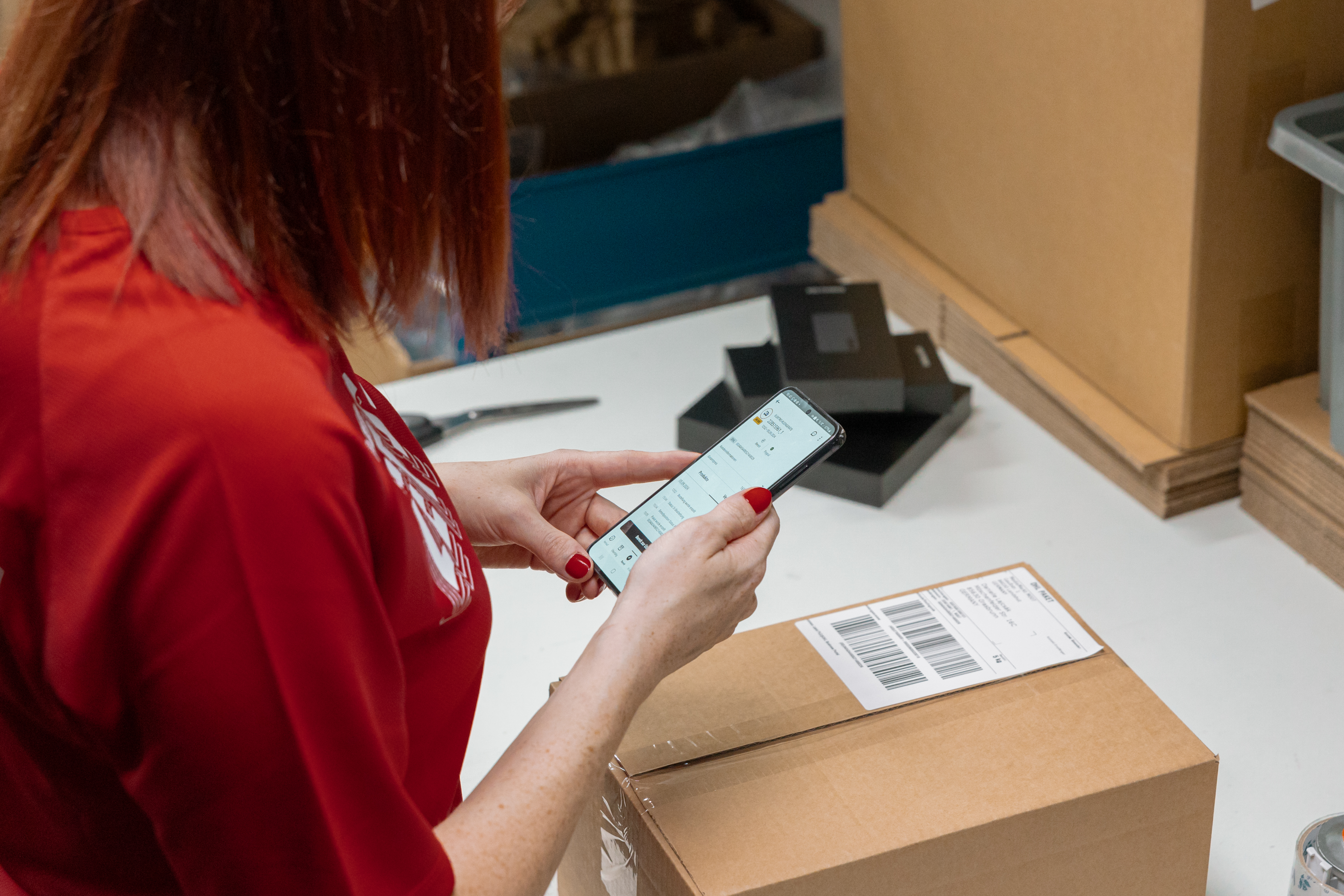 A Media Markt employee wearing a red shirt reads an order from their phone while standing in front of a table with a sealed cardboard box.